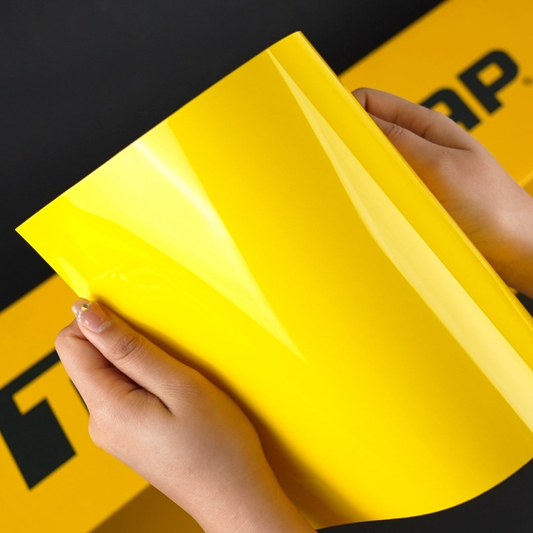 Close-up view of a person holding TeckWrap Sunray Yellow vinyl sheet, showcasing its glossy texture and rich, sunny hue.