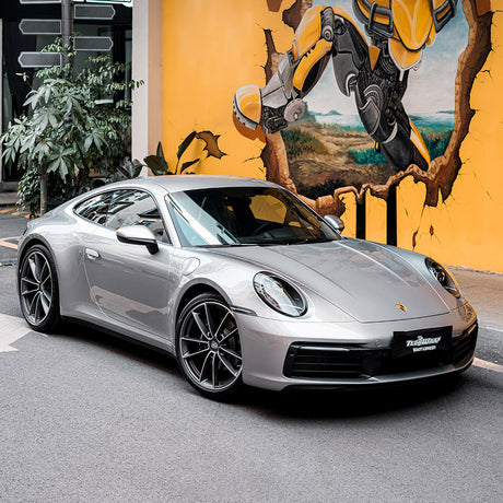 A full side view of a Porsche sports car wrapped in TeckWrap’s Dolomite Silver RB33-HD Gloss Vinyl Wrap, parked against a colorful graffiti wall, highlighting the clean, luxurious silver gloss finish on its entire body.