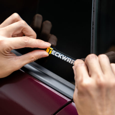 Hands applying TeckWrap 8cm logo sticker on car window for branding or decoration.