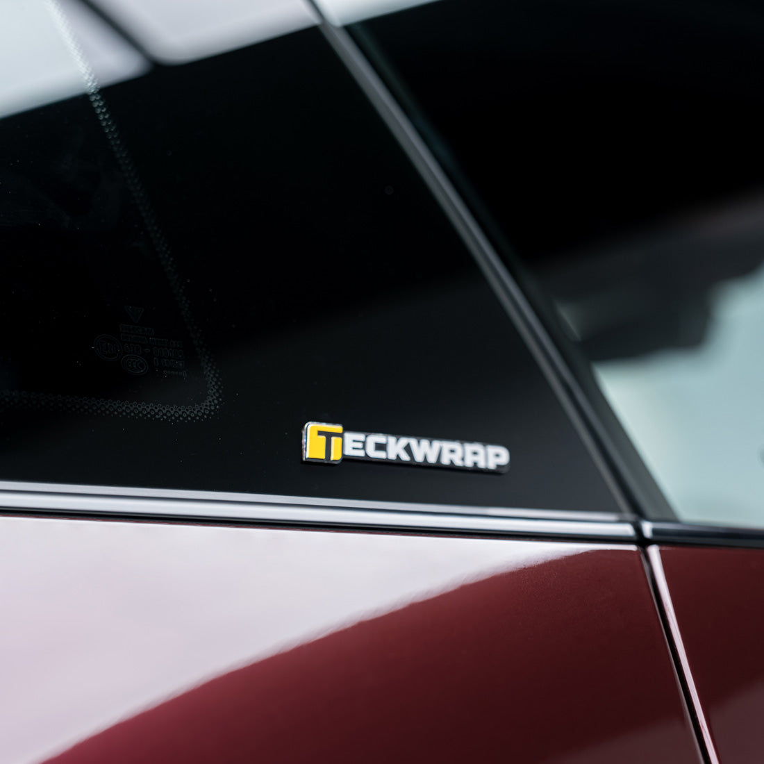 TeckWrap small logo decal applied on side window of maroon vehicle.