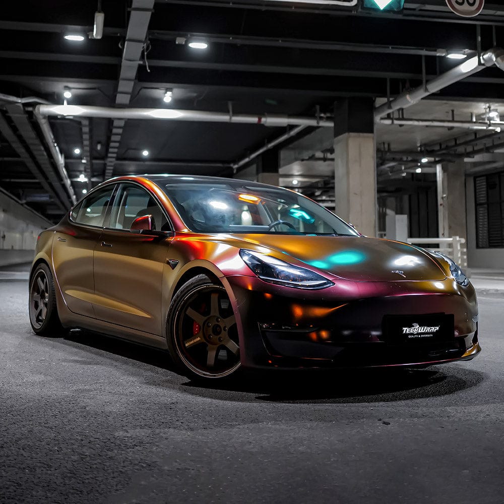 Front angled view of Tesla Model 3 in TeckWrap Madeira Red Matte chameleon vinyl wrap with a glowing, multitone matte finish under artificial lighting.