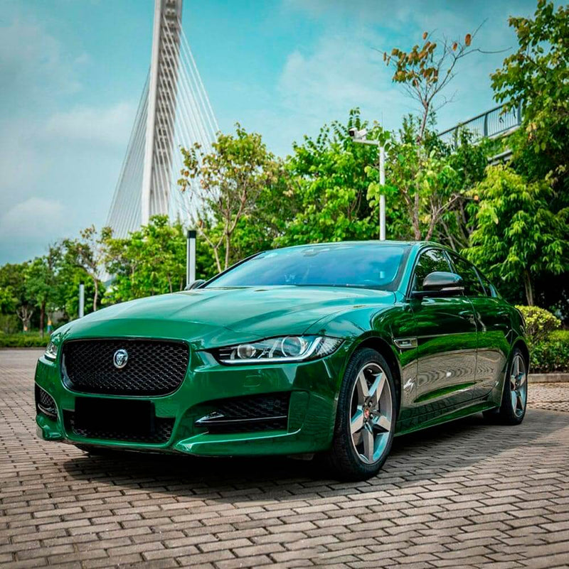 Elegant Jaguar sedan fully wrapped in TeckWrap Gloss Amphibian Green (CG32-HD), set against an urban greenery backdrop.