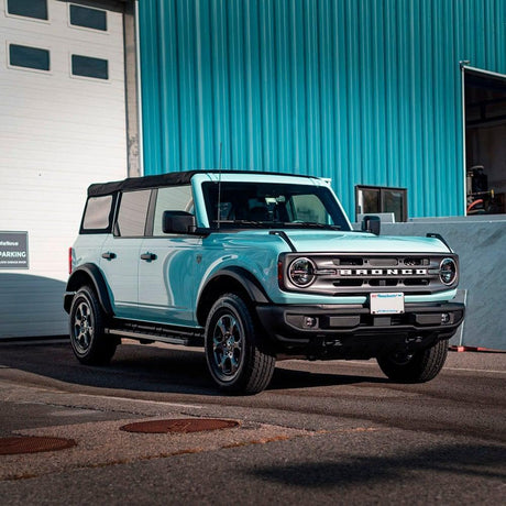 Ford Bronco wrapped in TeckWrap Gloss Bali Blue (CG37-HD) vinyl parked in front of a teal industrial building, showcasing the vibrant high-gloss blue finish in natural daylight.