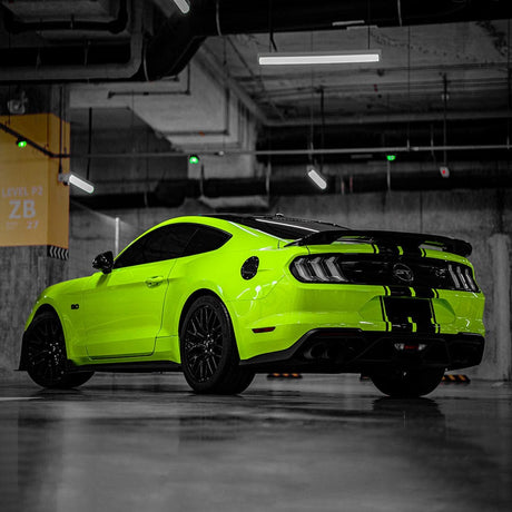Rear-side view of a Mustang wrapped in TeckWrap Gecko Green gloss vinyl, enhanced with striking black racing stripes, in a sleek indoor parking environment.