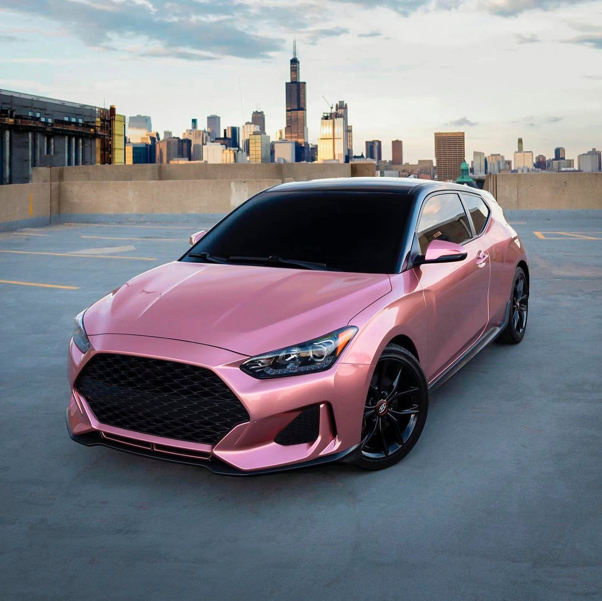 Hyundai Veloster sporting TeckWrap Pink Gold (GAL23-HD) Gloss Vinyl Wrap with a stunning cityscape background, accentuating its sophisticated, metallic appearance.