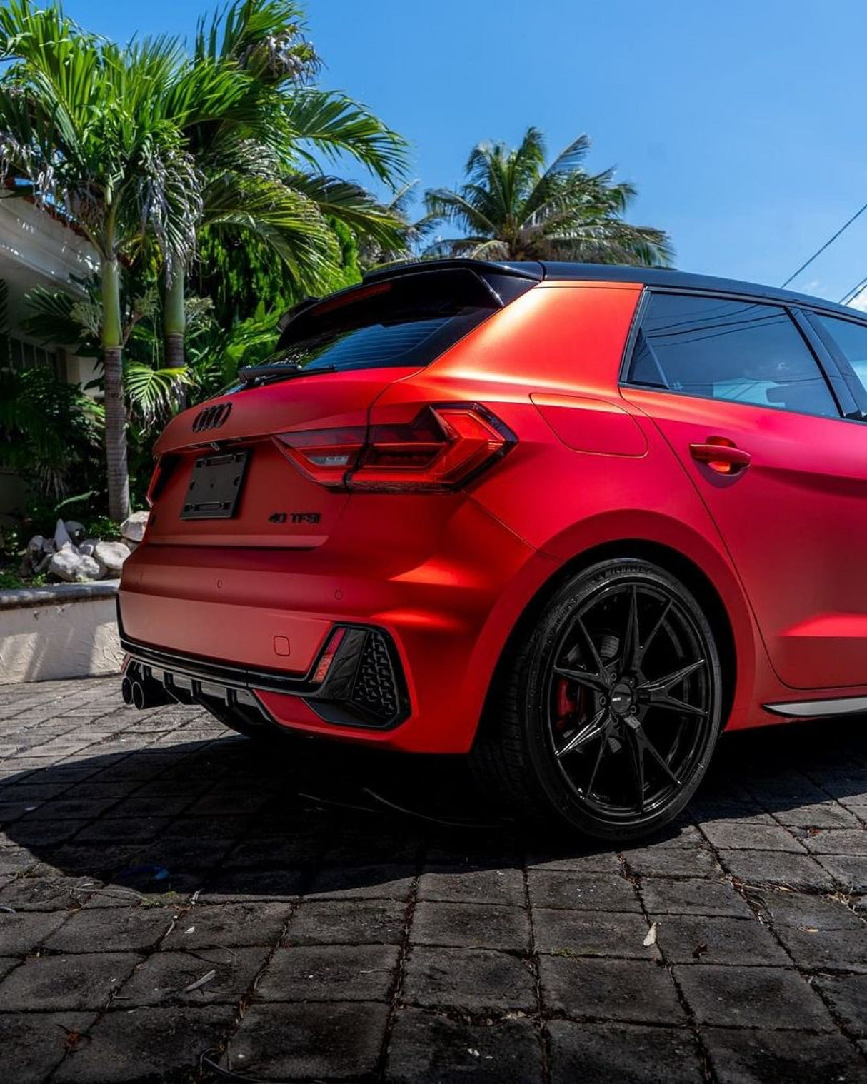 Rear quarter view of an Audi hatchback in TeckWrap Flame Red SMT15 satin vinyl wrap, parked in a lush, tropical setting.