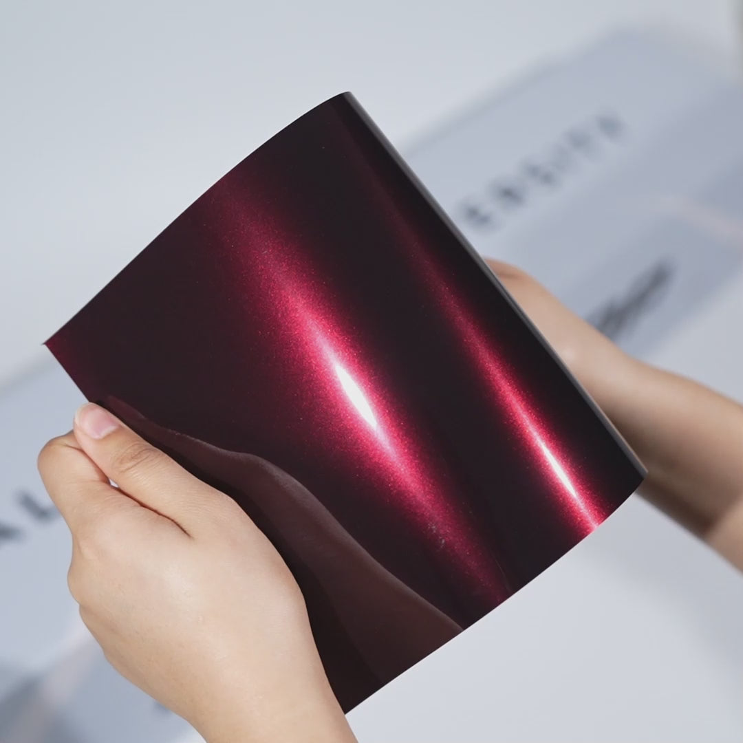 Hands holding a TeckWrap Deep Maroon Metallic PPF sample (CPX415), showcasing its rich wine-red metallic gloss and premium paint protection finish.