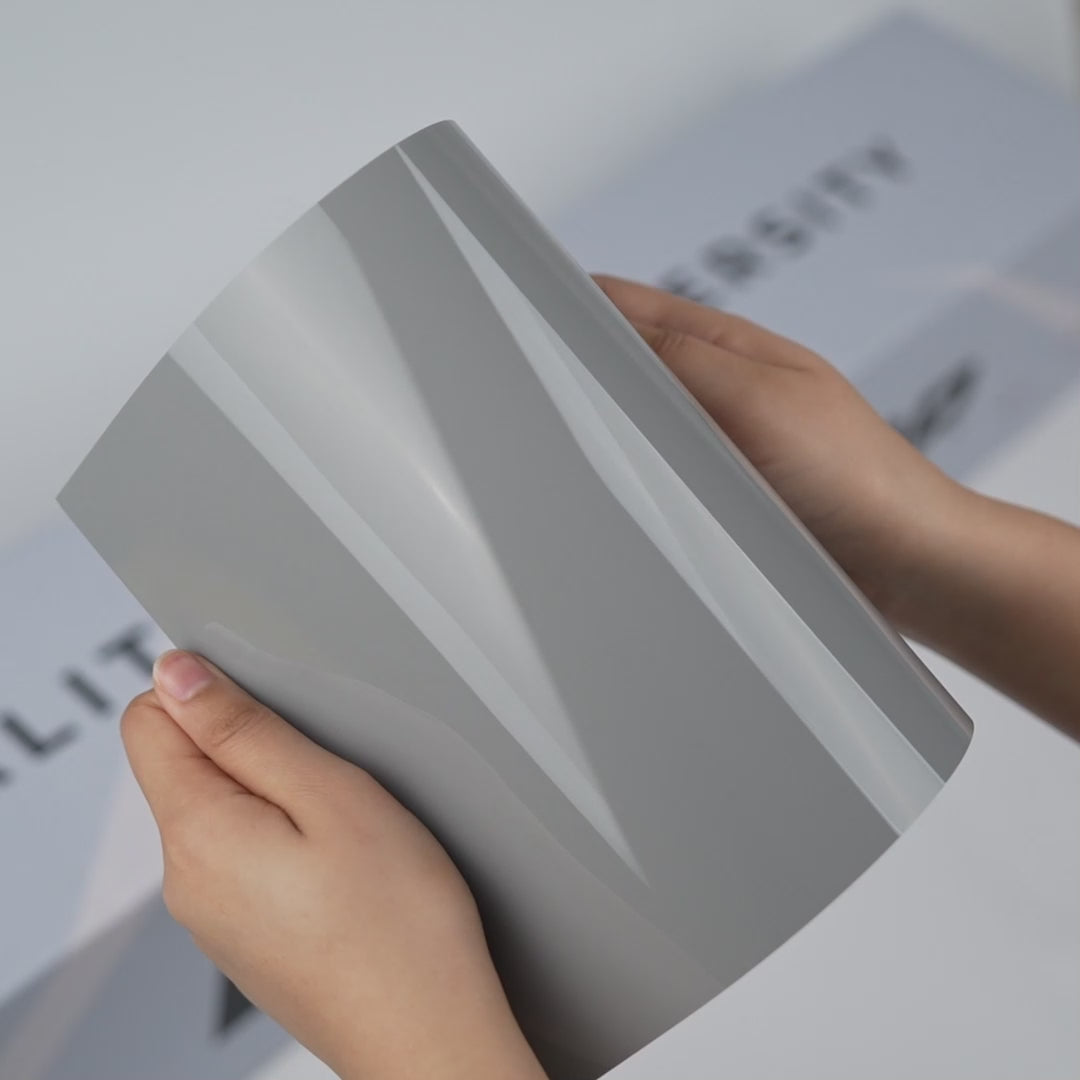 Hands holding a TeckWrap Stone Grey Gloss Color PPF sample (CPX307), showcasing its smooth glossy stone-grey finish and premium paint protection surface.