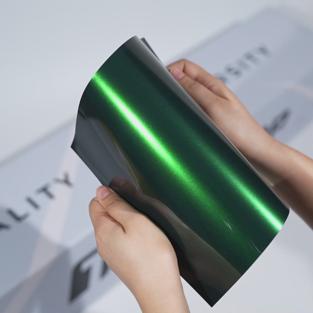 Hands holding a TeckWrap Noble Green Gloss Color PPF sample (CPX502), showcasing its brilliant gemstone-like green shine and premium paint protection finish.