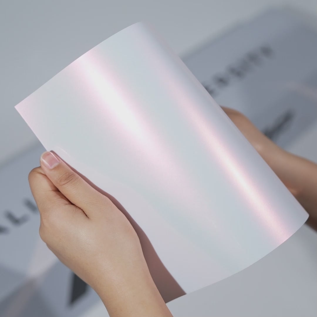 Hands holding a TeckWrap Sakura Pearl White PPF sample (CPX404), showcasing its glossy pearlescent white finish with a pink sheen and premium paint protection surface.