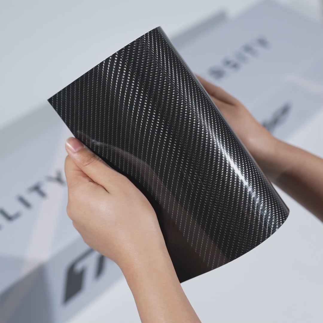 Hands holding a TeckWrap Sable Carbon Gloss PPF sample (CPX601), displaying the premium black carbon-fiber texture and high-gloss paint protection finish.