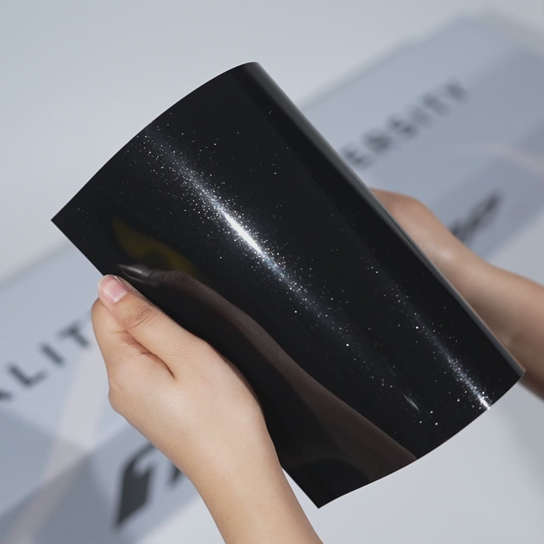 Hands holding a TeckWrap Luxe Black Metallic PPF sample (CPX409), highlighting its glossy black metallic sparkle and premium paint protection finish.