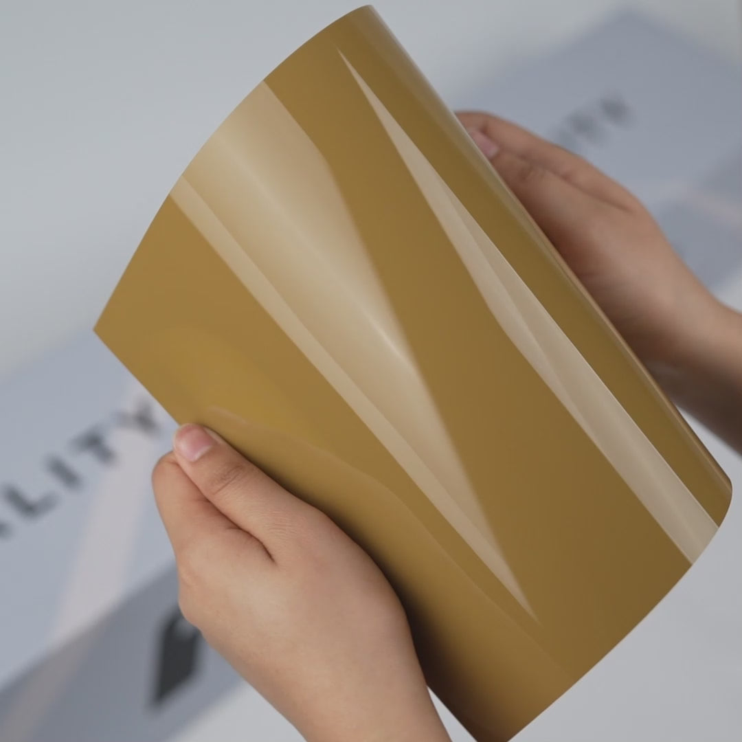 Hands holding a TeckWrap Golden Fleece Gloss Color PPF sample (CPX304), highlighting its warm glossy golden-beige finish and premium paint protection surface.