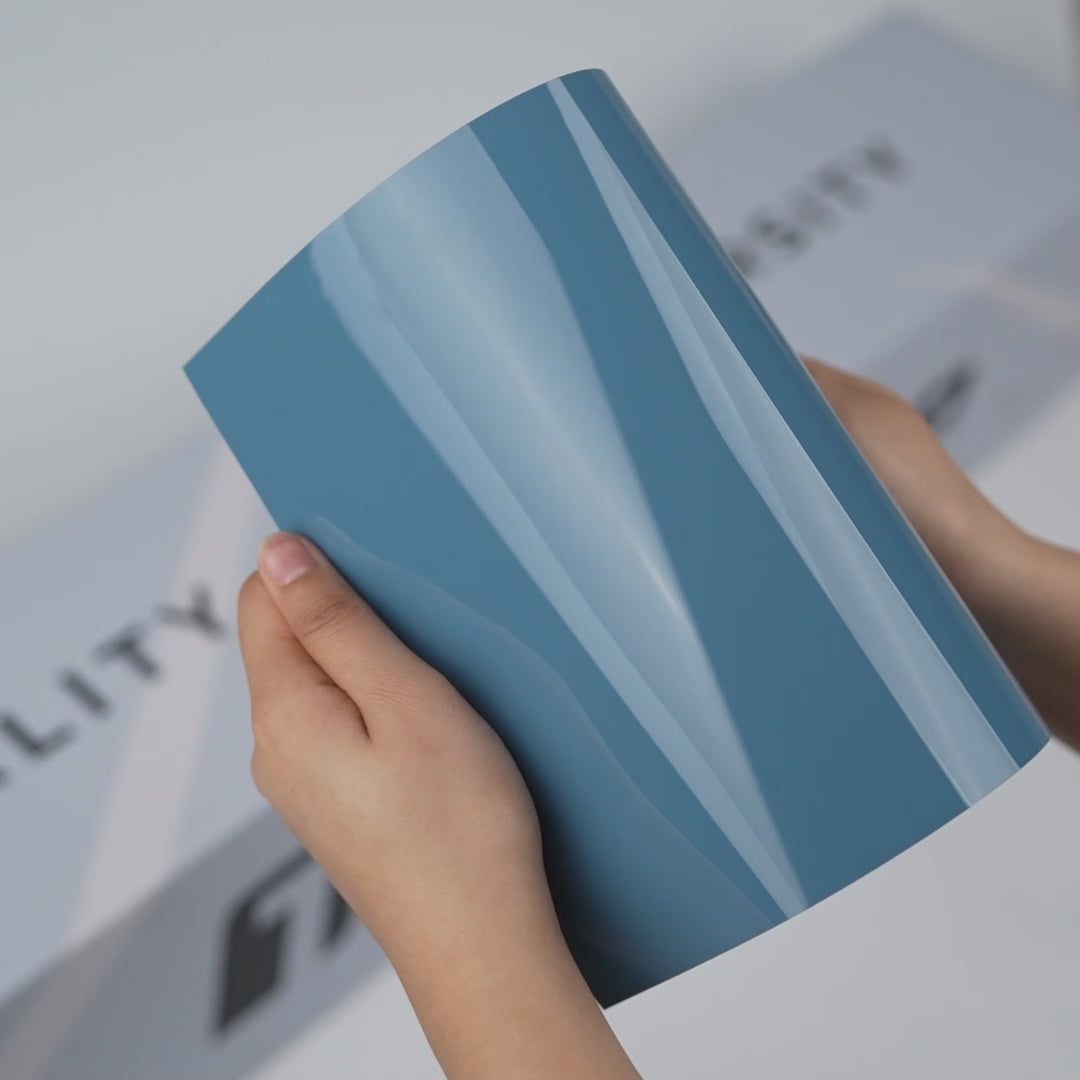 Hands holding a TeckWrap Porcelain Blue Gloss Color PPF sample (CPX310), showcasing its smooth glossy porcelain-blue finish and premium paint protection surface.