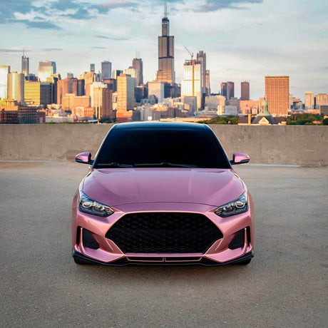 Front view of Hyundai Veloster wrapped in glossy TeckWrap Pink Gold (GAL23-HD) vinyl, highlighting its vibrant pink-gold metallic color against a dramatic urban skyline.