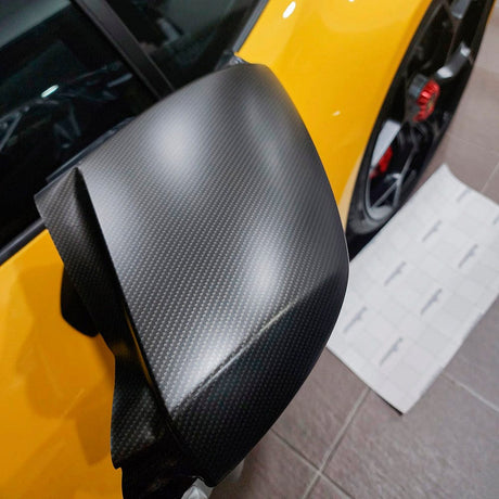 Installation process detail of a vehicle's side mirror being wrapped with TeckWrap Matte Ceramic Carbon (RCF03M) Vinyl Wrap, emphasizing its precision fit, flexibility, and attractive matte carbon texture.